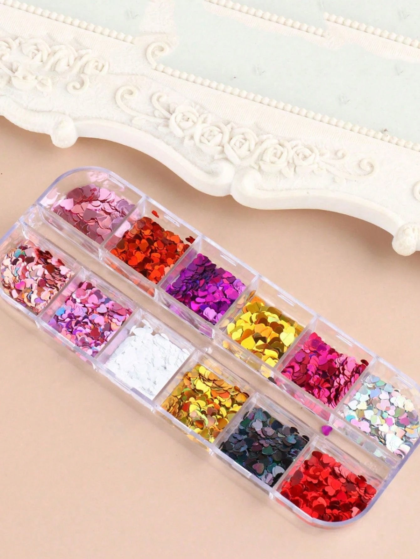 A clear plastic organizer tray filled with assorted decorative glitter flakes in multiple colors, including pink, red, purple, gold, silver, and dark blue, arranged on a light beige surface with a white ornate frame in the background.